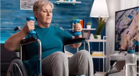 An elderly woman in a wheelchair exercises with blue dumbbells in a living room.