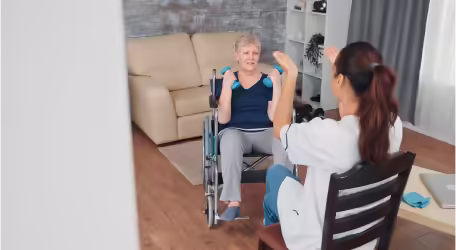 An elderly woman in a wheelchair exercises with blue dumbbells in a living room.