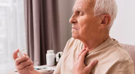 An elderly man with white hair holds a white inhaler to his nose, while his other hand rests on his chest/throat area, suggesting he is experiencing discomfort or breathing difficulties.
