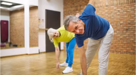 Two seniors, a man and a woman, performing exercises with outstretched arms in a bright fitness studio.
