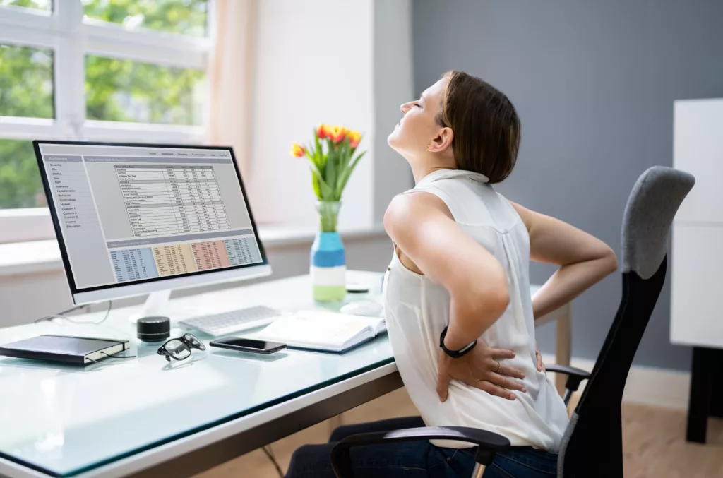 A female sitting in front of the computer table and looking annoyed with the back pain.